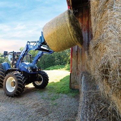 A blue tractor with a front loader is lifting a large round hay bale near a barn. The tractor is positioned on a gravel surface with a grassy field in the background, under a clear blue sky.