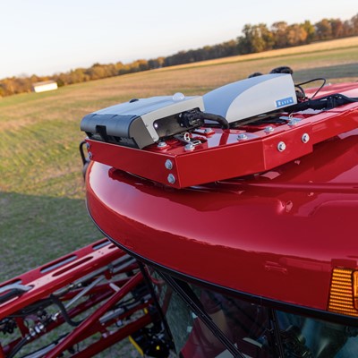 A close-up view of the top of a red agricultural machine, featuring mounted equipment including sensors and communication devices. The background shows a green field under a clear sky, indicating a rural farming environment.