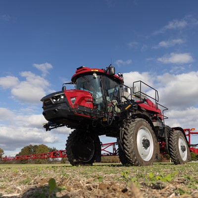 A red agricultural sprayer vehicle parked on a field under a partly cloudy sky. The sprayer features large wheels, a high cabin, and is equipped for crop spraying, showcasing its modern design and functionality.