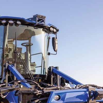 Close-up view of a modern blue agricultural tractor, showcasing its cab with large windows, advanced lighting, and various hydraulic components. The background features a clear blue sky, emphasizing the tractor's design and functionality for farming tasks.