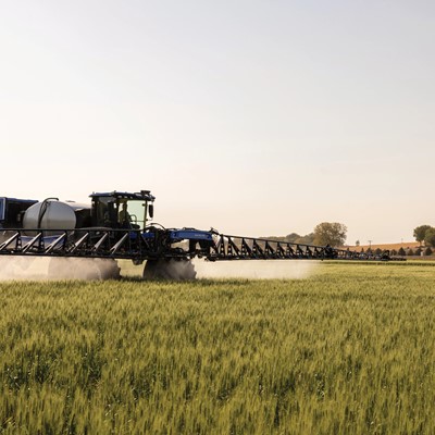 A blue agricultural tractor equipped with a large sprayer is working in a green field during a clear day. The tractor is spraying a mist over the crops, with trees and a clear sky in the background.