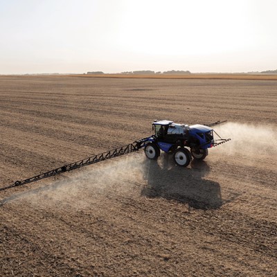 A blue tractor equipped with a sprayer is working in a vast, empty agricultural field. Dust is being kicked up as the tractor moves across the dry, brown soil under a clear sky. The landscape is flat and stretches into the distance, showcasing the agricultural setting.