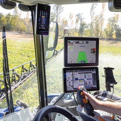 A close-up view of the interior of a modern agricultural tractor. The dashboard features multiple digital screens displaying various farming data and controls. A person’s hand is seen on the steering wheel, while the tractor is in a field with green crops in the background. The environment is bright and sunny, indicating a clear day for farming activities.