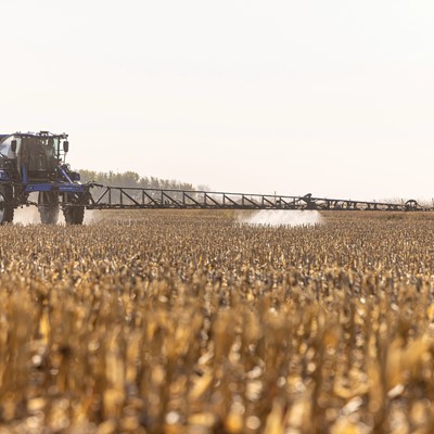 A large agricultural sprayer tractor applying chemicals to a field of dry, golden crops under a clear sky. The tractor is positioned in the distance, with a long spray arm extending outward, creating a mist as it moves through the harvested field.