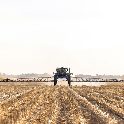 A large agricultural sprayer machine operates in a field, applying chemicals to the soil. The landscape features rows of harvested crops, with a clear sky in the background, indicating a sunny day.