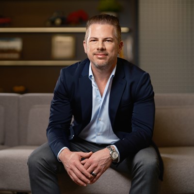 A professional man stands confidently in a modern office environment. He is wearing a dark blazer over a light blue shirt, with a slight smile on his face. The background features a blurred office space with plants and soft lighting, creating a welcoming atmosphere. The man is leaning against a wooden table, exuding a sense of professionalism and approachability.
