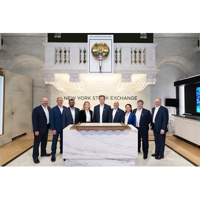 A group of nine business professionals standing behind a marble podium inside the New York Stock Exchange building, with the iconic NYSE bell and "New York Stock Exchange" sign visible on the wall behind them. The individuals are dressed in formal business attire, posing for a group photo in a well-lit, elegant interior space.