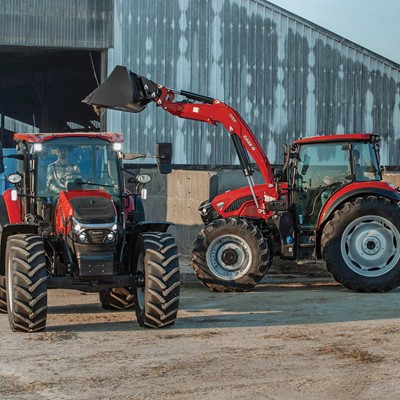 Two red tractors are positioned in front of a large barn with a corrugated metal exterior. One tractor is equipped with a front loader lifting a bucket filled with material, while the other tractor is attached to a blue trailer. The scene appears to be on a farm with dirt ground and hay visible near the barn.
