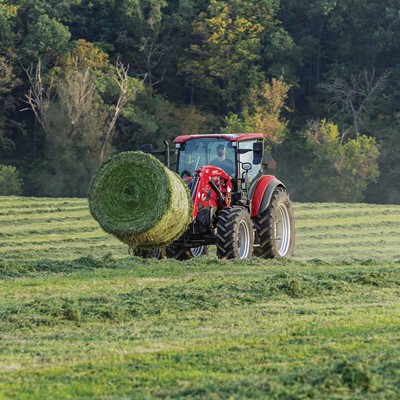 A red tractor is carrying a large round bale of freshly cut green hay in a vast field with rows of cut grass, surrounded by dense trees with green and autumn-colored leaves in the background.