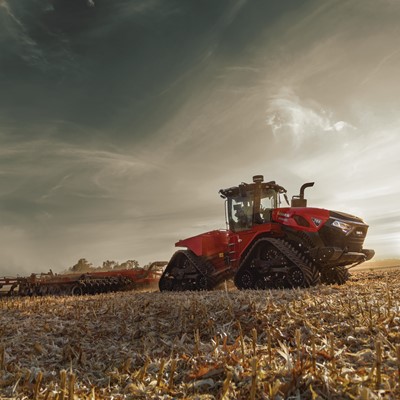 A large red tracked tractor working in a harvested field during sunset, pulling farming equipment to till the soil under a dramatic cloudy sky.