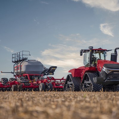 A large red Case IH agricultural tractor with rubber tracks is pulling a wide, complex farming implement across a dry field under a partly cloudy sky during daylight.