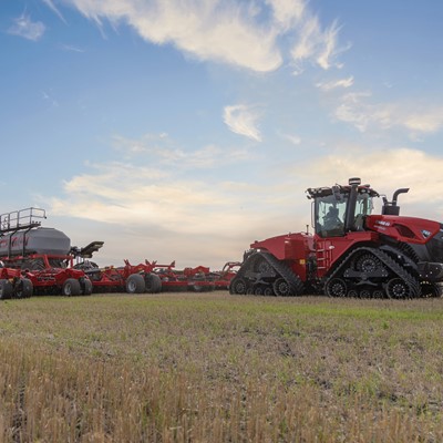 A large red Case IH tractor with rubber tracks is pulling a wide agricultural planter across a field under a partly cloudy sky during sunset or sunrise. The field has short stubble and green patches, indicating recent harvesting or planting activity.