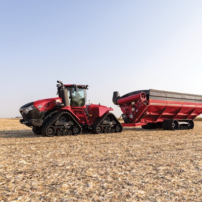 A large red agricultural tractor with rubber tracks is parked in a harvested field, attached to a matching red grain cart with rubber tracks under a clear blue sky.