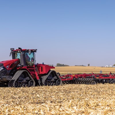 A large red Case IH tractor with rubber tracks is pulling a wide cultivator implement across a harvested cornfield under a clear blue sky, preparing the soil for planting.