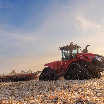 A large red Case IH tractor with rubber tracks is working in a harvested field at sunset, pulling farming equipment behind it under a partly cloudy sky.