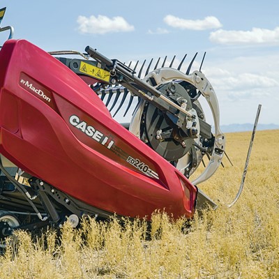 A close-up view of a red Case IH FD240 agricultural harvesting machine by MacDon operating in a golden crop field under a partly cloudy blue sky. The machine's cutting and gathering components are visible, with dry crops surrounding it.