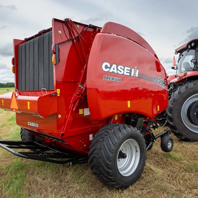 A red Case IH RB566 Premium round baler attached to a red tractor, positioned on a grassy field with hay bales scattered in the background under a cloudy sky.