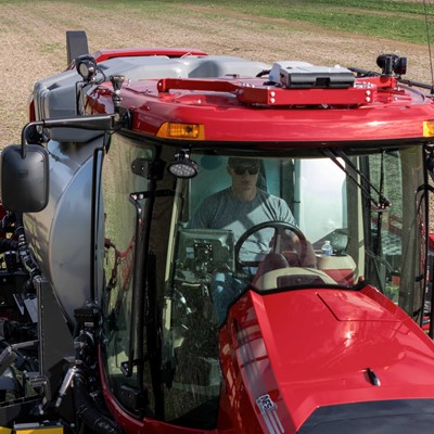 A man wearing sunglasses operates a large red agricultural tractor with a sprayer attachment in a field with sparse green crops and dry soil. The tractor has a spacious glass cabin and large rear tires, and the man is seated inside, focused on driving.