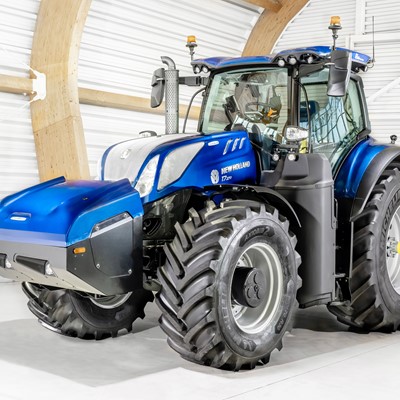 A blue New Holland T7.270 tractor with large black tires is parked inside a bright, modern barn with white walls and wooden beams. The tractor features a spacious enclosed cabin and front weights for balance.