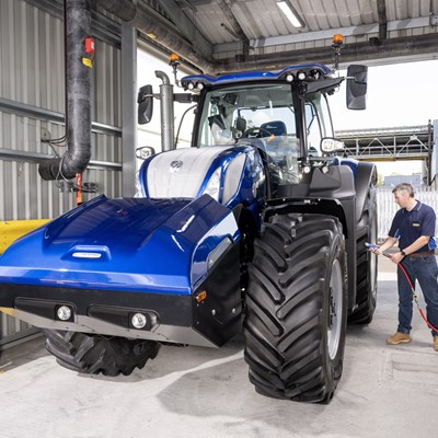 A man in a blue shirt and jeans is inspecting or charging a large blue agricultural tractor inside a modern industrial facility with metal walls and safety barriers.