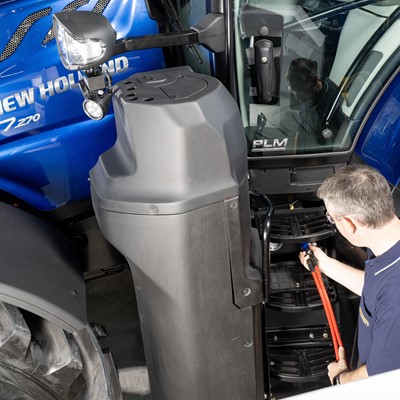 A man in a navy blue shirt is inspecting or repairing the steps of a blue New Holland T7.270 tractor inside a workshop or garage. The tractor's large front tire and side mirror are visible, along with the "Methane Power" label on the tractor's hood.