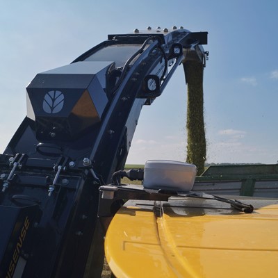 A close-up view of a modern agricultural harvester unloading freshly harvested crops through a chute into a trailer on a sunny day, with a clear blue sky and green fields in the background.