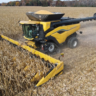 A yellow combine harvester is actively harvesting a large cornfield, with dry corn stalks being cut and collected. The machine's grain tank is partially filled with harvested corn, and dust rises from the field as the harvester moves forward. Trees with autumn foliage are visible in the background under a clear sky.