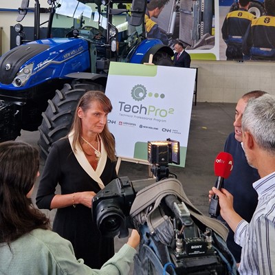 A woman is being interviewed by a news crew inside a workshop or showroom with a blue New Holland tractor in the background. Behind her is a large sign displaying the "TechPro2" logo and the text "Technical Professional Program." The setting includes posters of people working on machinery, emphasizing a technical or agricultural theme. The interviewers hold microphones and a professional video camera is recording the scene.