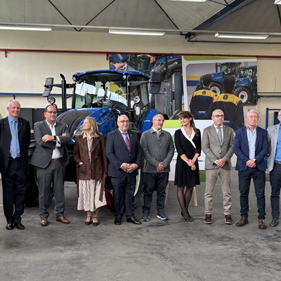 A group of twelve professionally dressed people standing in a row inside an industrial workshop with a large blue tractor behind them. The setting appears to be a formal event or visit, with a mix of men and women posing for a group photo. The background includes industrial equipment, posters, and workshop lighting.