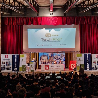 A large audience seated in an auditorium facing a stage with red curtains. On the stage, a speaker stands at a podium with a microphone, presenting in front of a large screen displaying the "TechPro² Technical Training Program" logo. Banners for Salesianos, New Holland, and CNH Industrial flank the stage. The event appears to be a formal presentation or training session.
