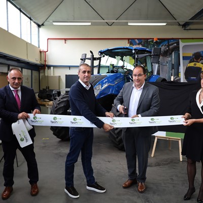 Five people standing in a workshop with a blue tractor in the background, participating in a ribbon-cutting ceremony. One man in a gray suit is cutting a white ribbon with "TechPro" logos, while the others hold the ribbon and smile. The setting appears to be an industrial or agricultural equipment facility.