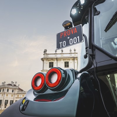 Close-up view of the rear part of a modern black tractor with red circular tail lights and a license plate reading "PROVA TO 001," set against the backdrop of a historic European-style building and a cloudy sky at dusk.
