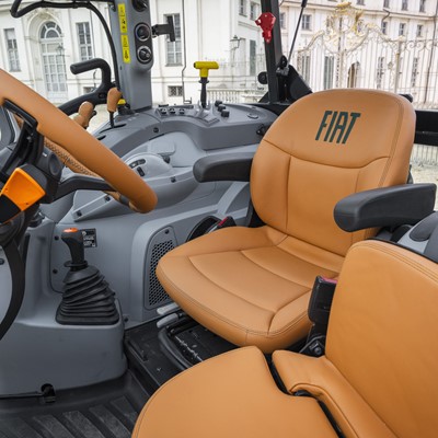 Interior view of a Fiat tractor cabin featuring tan leather seats, a steering wheel with an orange accent, various control levers, and a modern dashboard, with a building visible through the windows in the background.