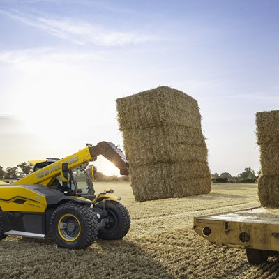 A yellow New Holland telehandler lifting large rectangular hay bales in a harvested field during sunset, with a flatbed trailer nearby stacked with more hay bales.