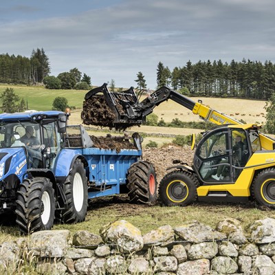 A blue tractor with a trailer is being loaded with soil or manure by a yellow New Holland telehandler in a rural field. The background features green rolling hills, trees, and a stone wall in the foreground under a cloudy sky.