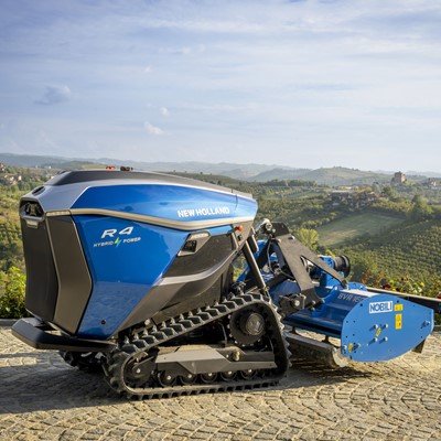 A blue New Holland R4 Hybrid Power autonomous agricultural robot with caterpillar tracks is parked on a cobblestone surface, with a scenic background of rolling green hills, vineyards, and a partly cloudy sky.