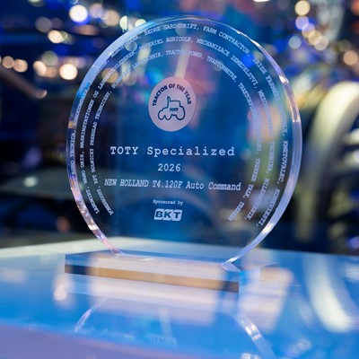 A clear circular glass award trophy with the inscription "TOTY Specialized 2026 NEW HOLLAND T4.120F Auto Command" and the logo "Tractor of the Year" featuring a tractor icon. The award is sponsored by BKT and is displayed on a reflective surface with a blurred background of blue and purple lights.