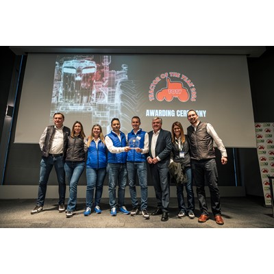 A group of seven people standing on a stage during the "Tractor of the Year 2026" awarding ceremony. They are posing for a photo, with two men in the center holding a transparent award plaque. Behind them is a large screen displaying a technical drawing of a tractor and the event logo featuring a red tractor silhouette with the text "TRACTOR OF THE YEAR 2026" and "AWARDING CEREMONY." The group is dressed casually, with some wearing blue vests and others in jackets and jeans.