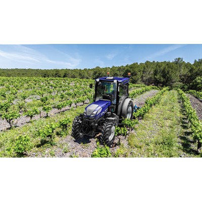A blue tractor is driving through a vineyard with rows of green grapevines under a clear blue sky, surrounded by a forest in the background.