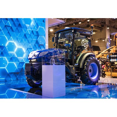 A modern blue agricultural tractor displayed indoors at an exhibition, featuring large rear tires, a glass-enclosed cabin, and illuminated by bright lights with a blue geometric patterned backdrop. A transparent display case with an award or trophy is positioned in front of the tractor.