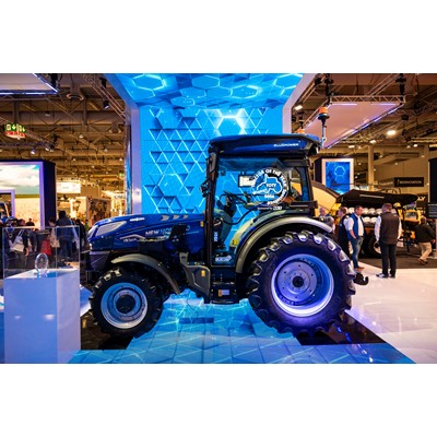 A blue New Holland tractor displayed at an indoor exhibition with a futuristic blue geometric backdrop. The tractor has large rear tires and smaller front tires, with a "Tractor of the Year 2026" award sticker on the cabin window. Several people are seen in the background, observing other exhibits. The setting is brightly lit with a modern, high-tech atmosphere.
