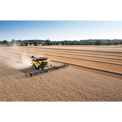 A yellow combine harvester is working in a vast golden wheat field, cutting and collecting the crop. The machine is moving through the field, leaving behind neat rows of harvested wheat. In the background, there are trees and a clear sky, indicating a bright and sunny day.