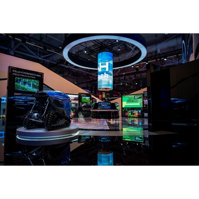 Exhibition booth showcasing advanced agricultural technology with a blue autonomous robotic vehicle on a circular platform, multiple digital screens displaying field preparation and seed planting information, and a large cylindrical hanging digital display with the word "Tech" in a modern indoor event space.