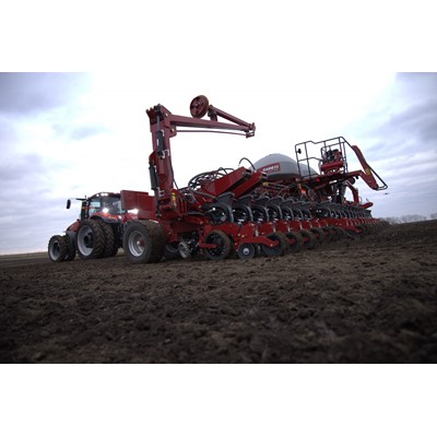 A large red agricultural tractor attached to a multi-row planter machine working on a freshly tilled field under a cloudy sky, preparing the soil for planting crops.
