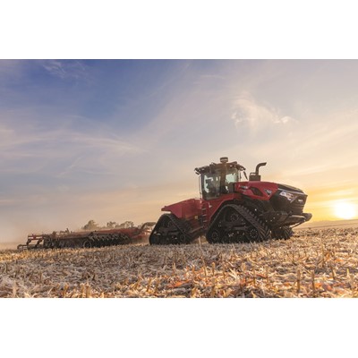 A large red Case IH tractor with rubber tracks is working in a harvested field during sunset, pulling farming equipment behind it under a partly cloudy sky.