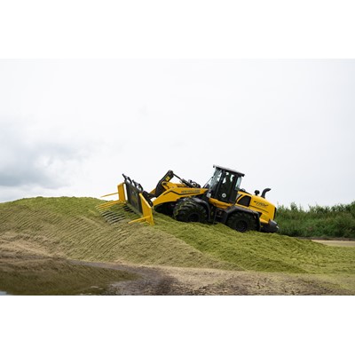 A yellow New Holland W170D wheel loader is pushing and leveling a large pile of green silage on a farm, with a cloudy sky in the background.