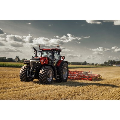 A red Case IH tractor with large tires is working in a golden wheat field under a partly cloudy sky, pulling a red agricultural implement for tilling the soil, with green crops and trees visible in the background.