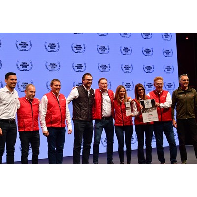 A group of nine people standing on stage in front of a large screen displaying the "Farm Machine 2026" logo repeatedly. Most individuals are wearing red vests over white shirts, while two are in different colored jackets. Two women in the center are holding awards, one holding a plaque and the other a certificate. The group is smiling and posing for a photo, suggesting a celebratory or award event.
