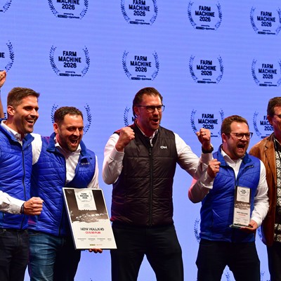 A group of six people standing on stage celebrating an award. Four men in blue and black vests are holding plaques and certificates, smiling and raising their fists in excitement. A woman on the left holds a microphone and a man on the right wears a brown leather jacket and patterned shirt. The background displays multiple "Farm Machine 2026" logos.