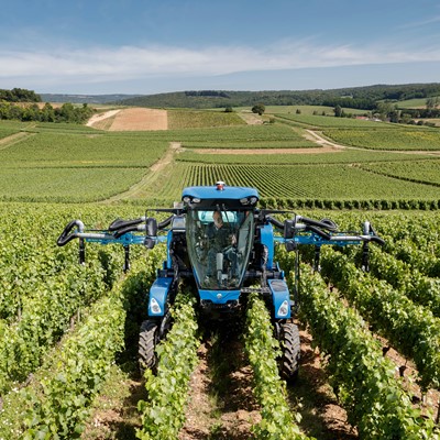 A blue agricultural machine is operating in a vineyard with rows of green grapevines under a clear blue sky, surrounded by rolling hills and farmland in the background.